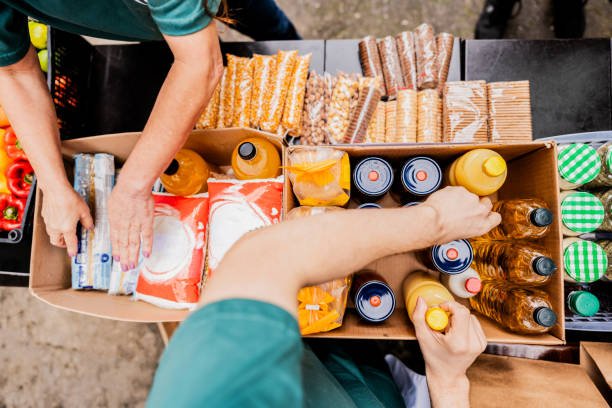 Two people reaching into a large cardboard box filled with bottled drinks, juice boxes, and snacks, with some snacks and bottles on the table nearby.