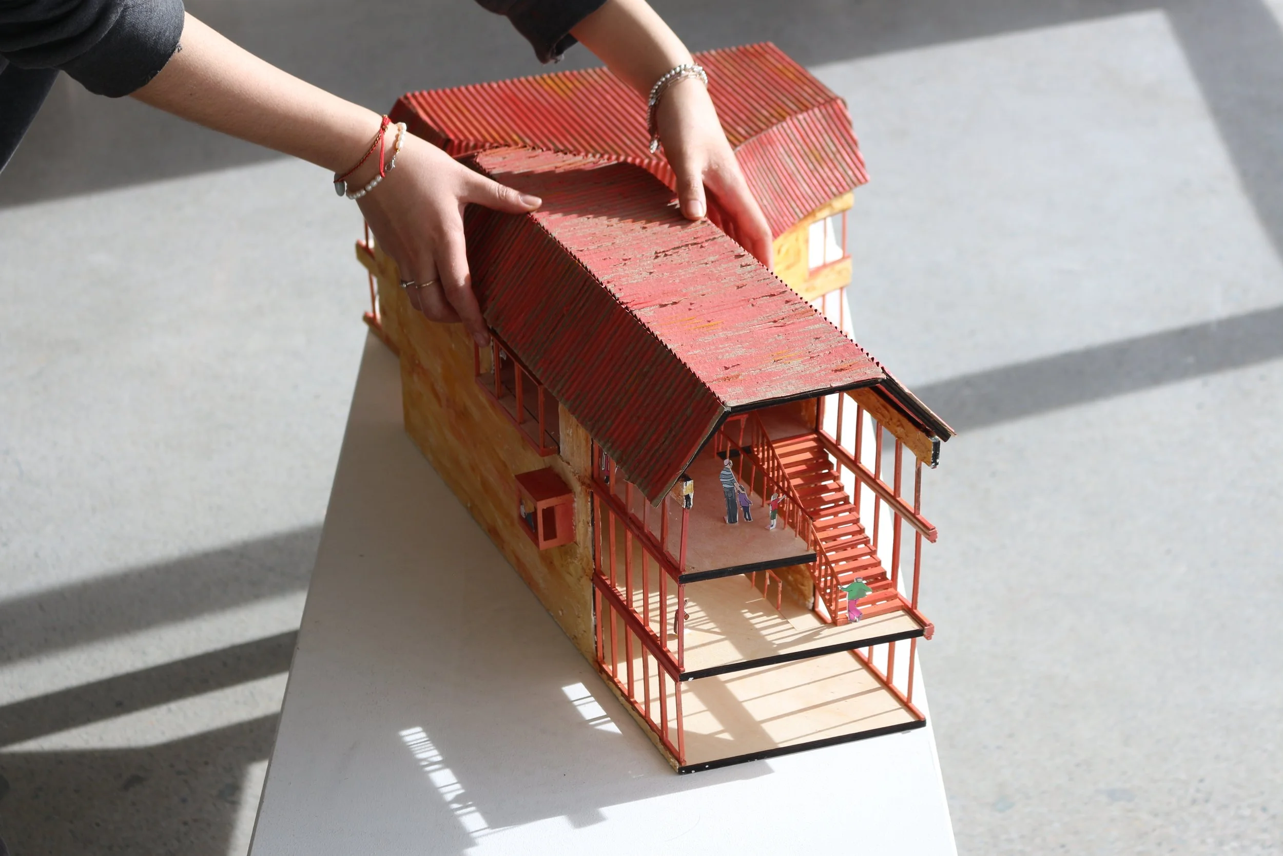 A detailed architectural model of a two-story building with a red roof and orange walls, featuring multiple levels, stairs, and small human figurines inside and outside.