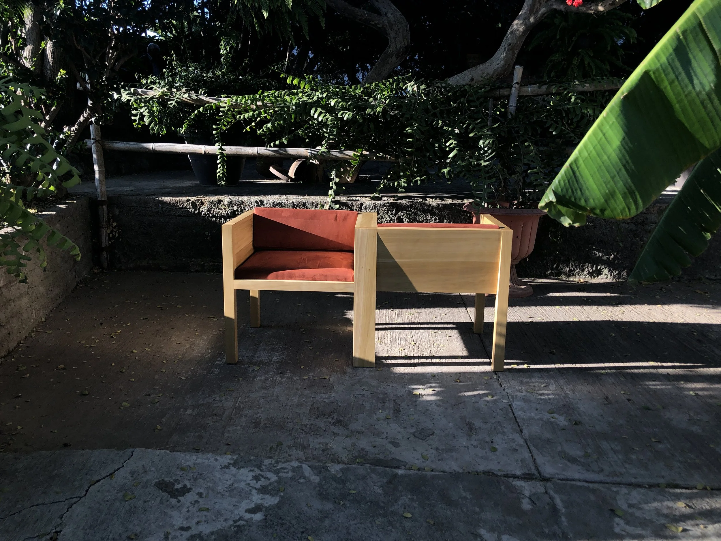 Banco de madera Yo - Tú en un espacio al aire libre rodeado de plantas y sombra.