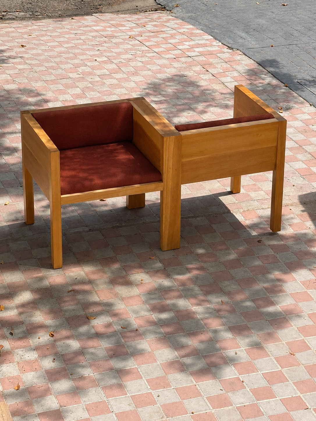 Sillón Yo -Tú para dos. Banca de madera con cojines de color rojo en una acera de baldosas, con sombra y algunas hojas secas.