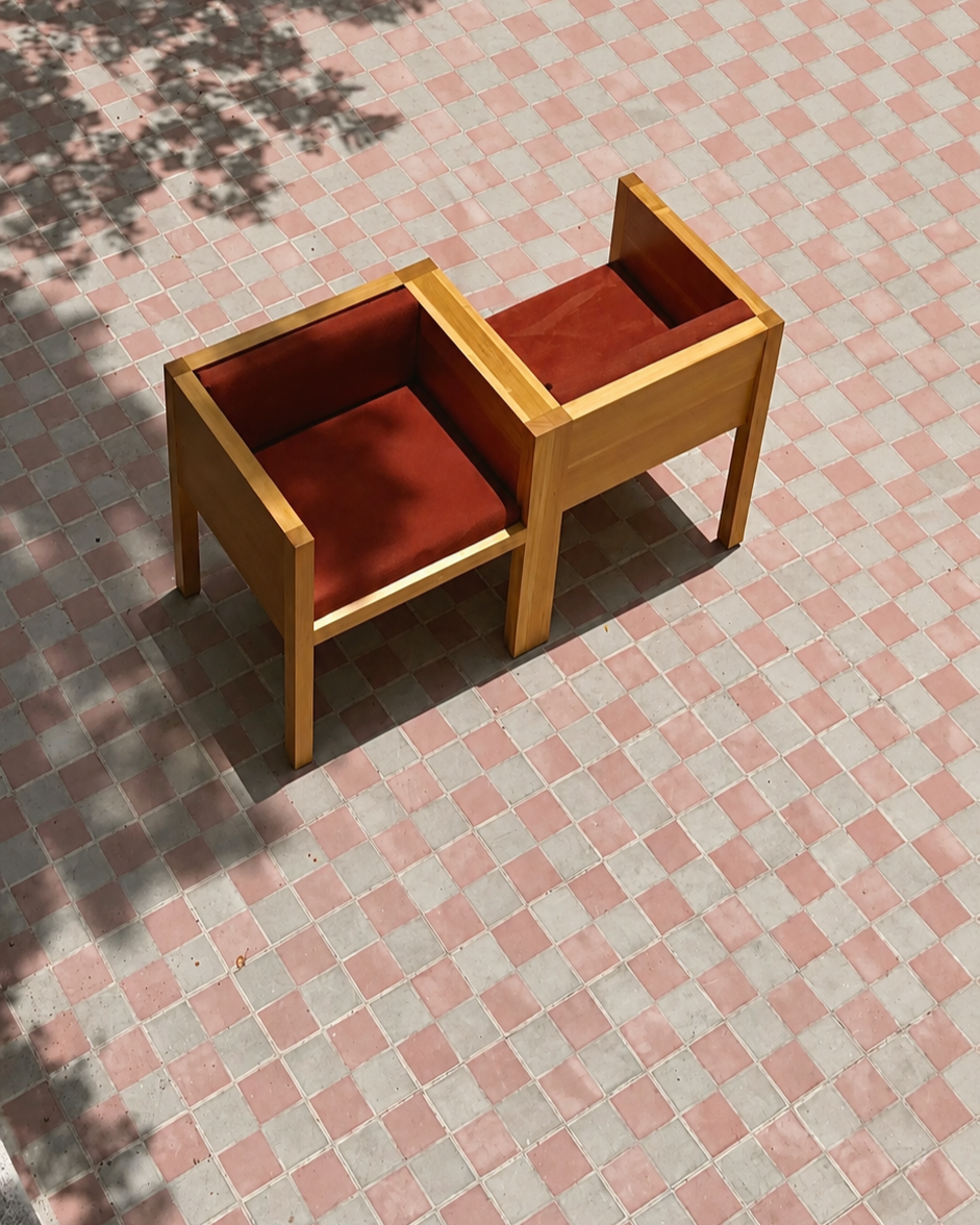 You - I lounge chair for two in oiled oak wood with red cushions is placed on a pink and gray traditional tiled outdoor surface in Guadalajara. Shaded shadows are cast across the tiles.
