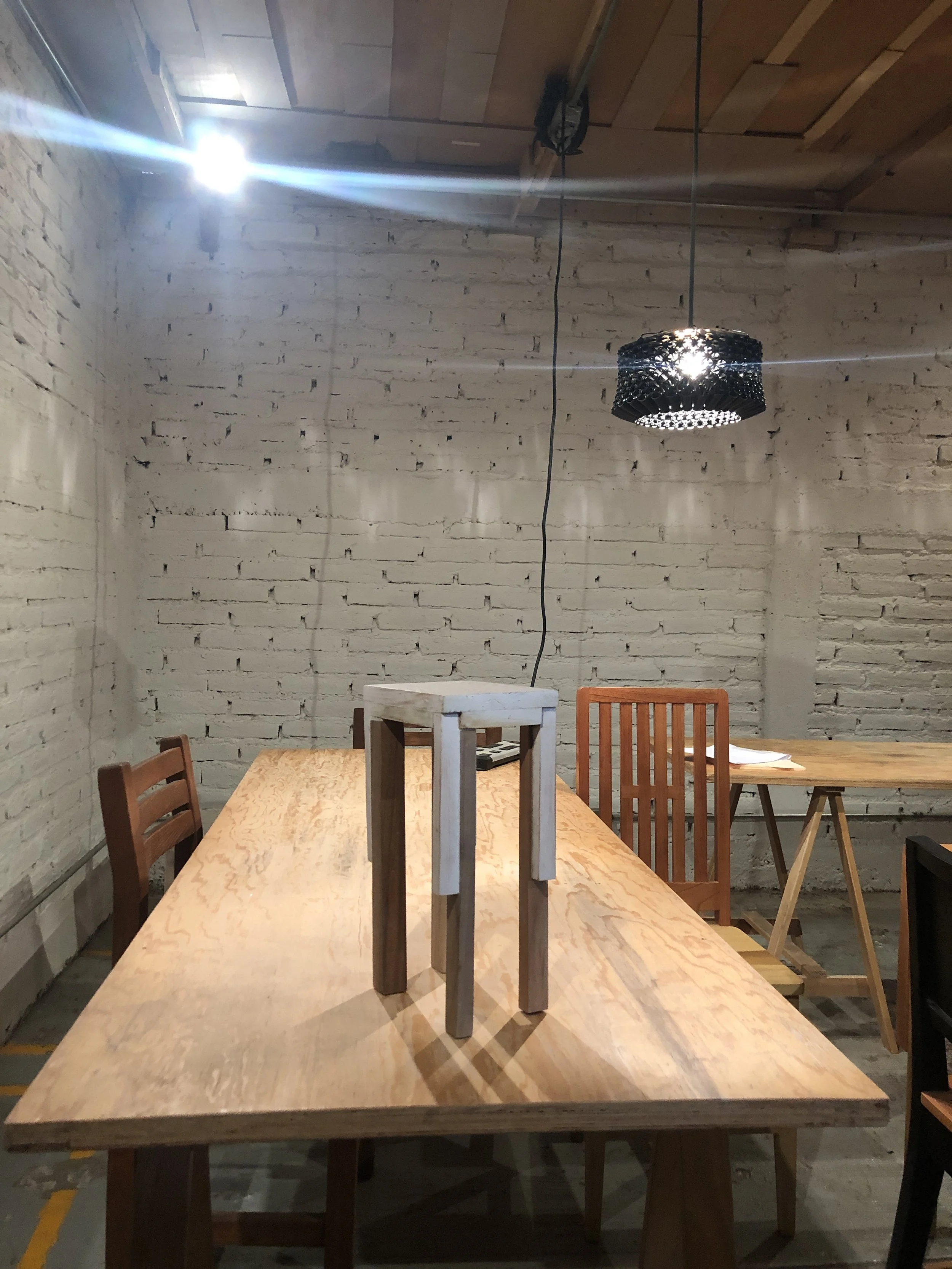 Vista de una tarifa de madera en un espacio cerrado con paredes de ladrillos pintados blancos, una lámpara negra con diseño de anillos de metal, y varias sillas de madera repartidas alrededor de la mesa.