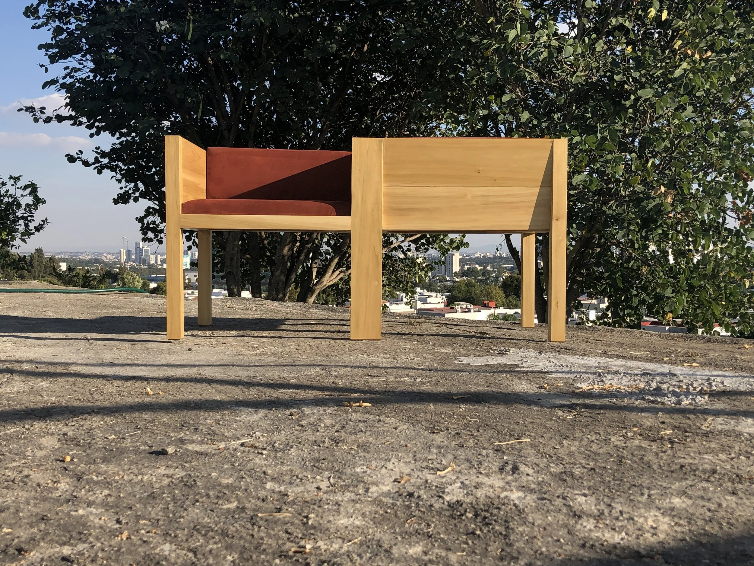 Sillón Yo -Tú para dos. Una estructura de madera con un asiento de color burdeos, situada al aire libre con fondo de árboles y ciudad en la distancia.