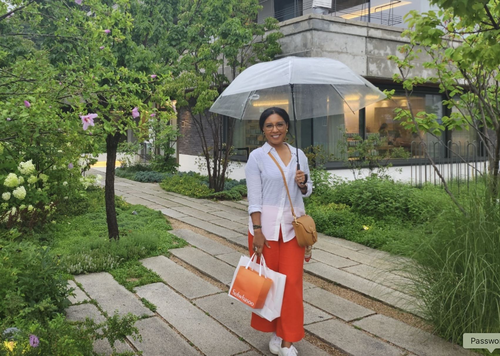 A smiling woman holding a transparent umbrella in a lush green garden with a stone pathway. She carries shopping bags labeled 'Sulwhasoo,' wears glasses, a striped shirt, red pants, and a tan crossbody bag Emily Maria Studios