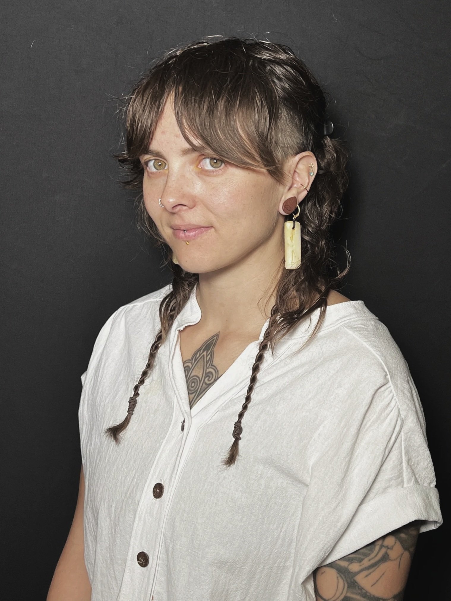 A young woman with short brown hair styled in braids, wearing a white button-up shirt with rolled-up sleeves, earrings, and a nose ring, posing against a black background.