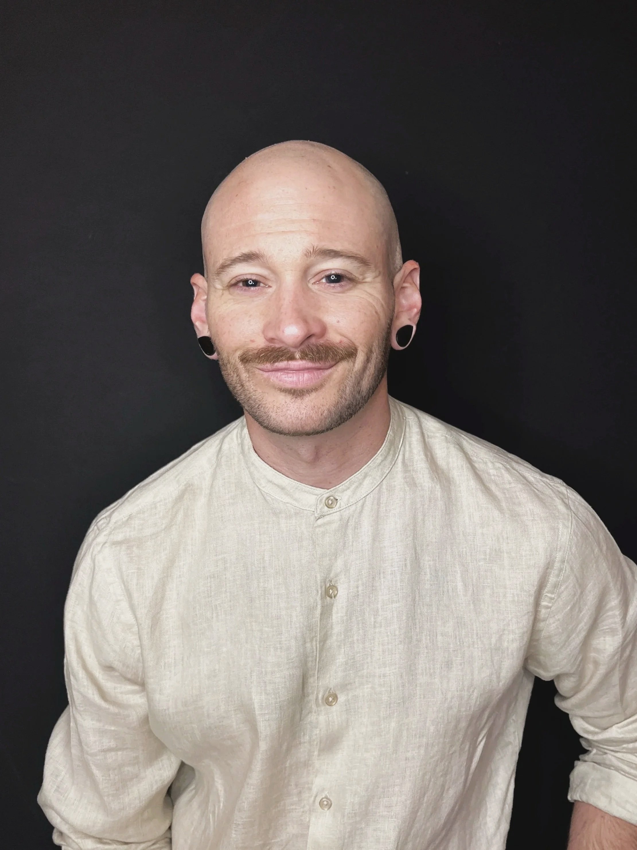 A bald man with pierced ears and a beard, wearing a beige button-up shirt, smiling against a black background.