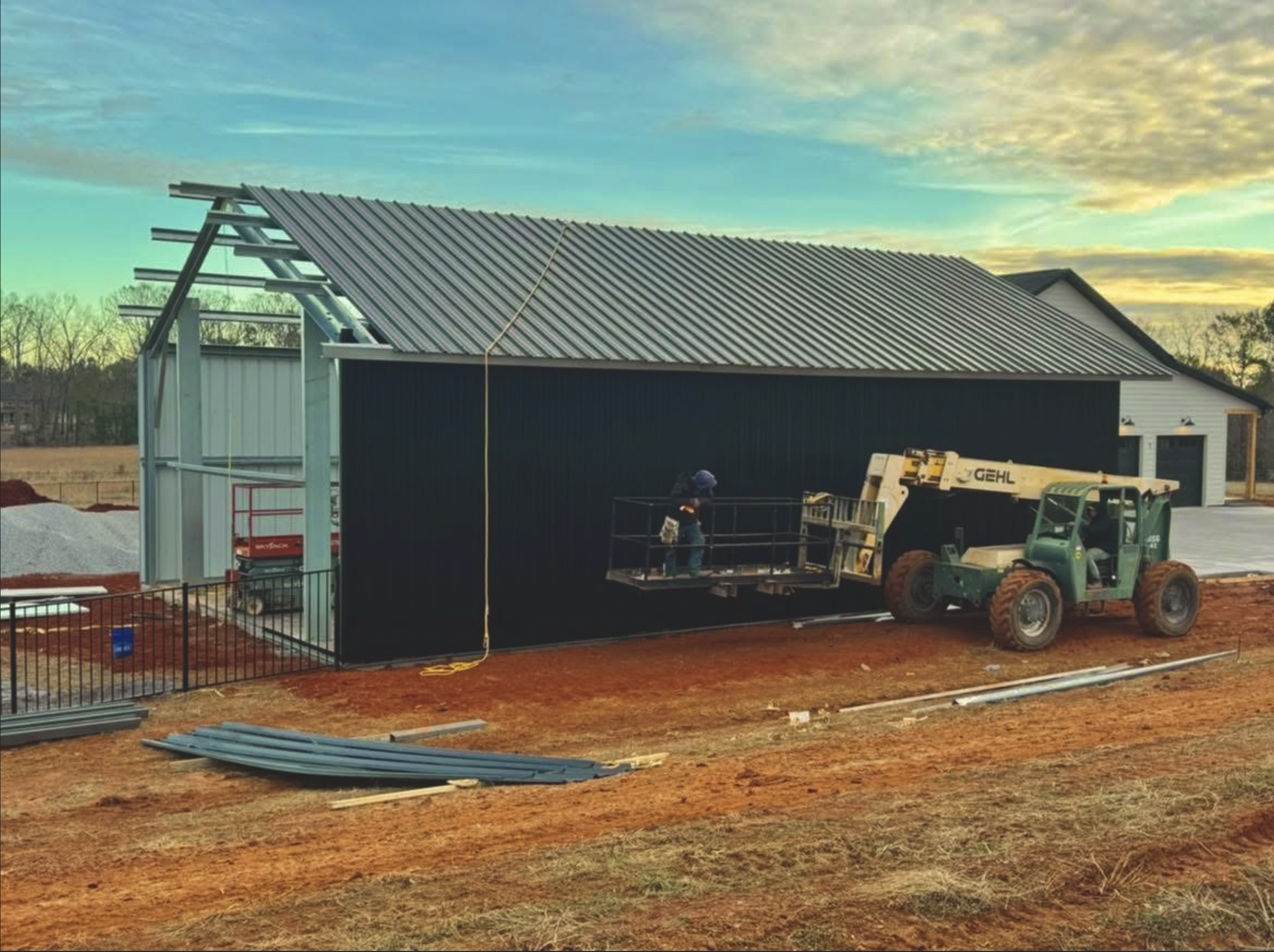 House under construction with a metal roof in progress, construction equipment and workers at the site during sunset.