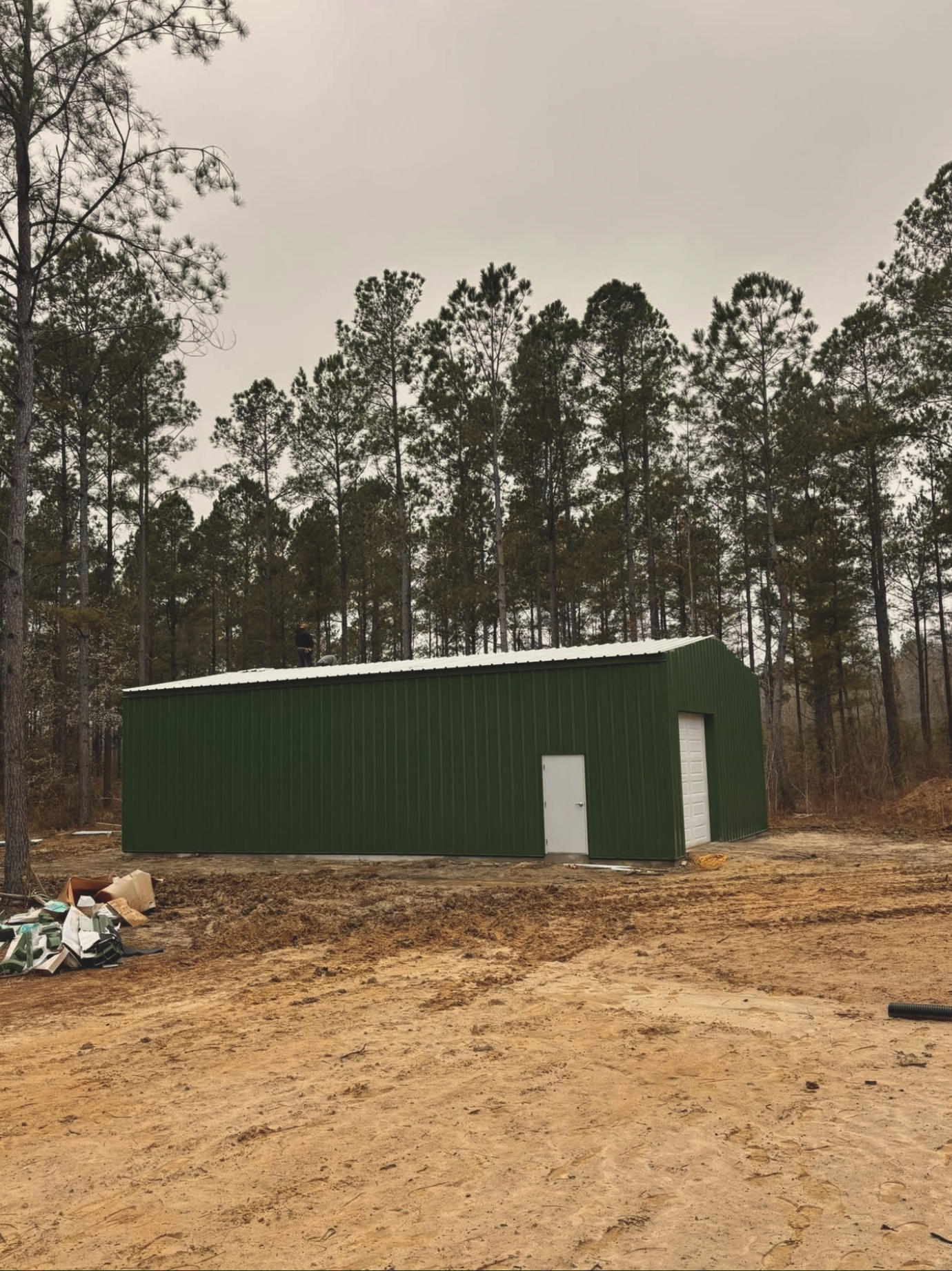 A green metal building with two white doors, situated on an uneven dirt ground area with some debris, against a backdrop of tall pine trees under an overcast sky.