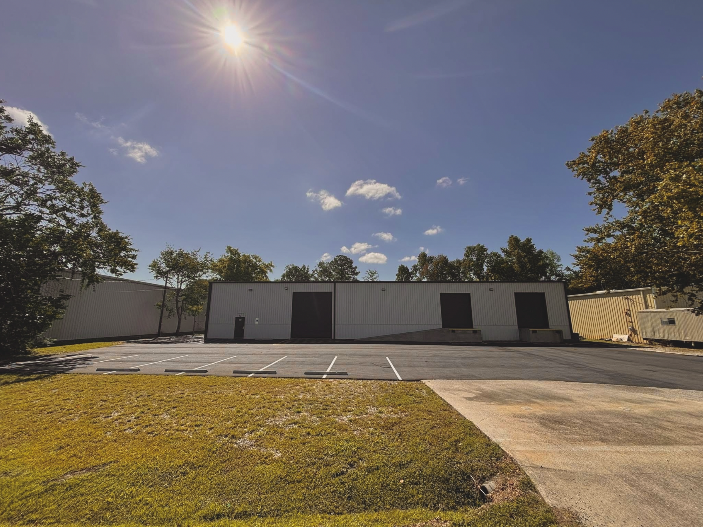 A parking lot in front of industrial or warehouse buildings with trees on either side and a clear blue sky with a few clouds and the sun shining brightly.