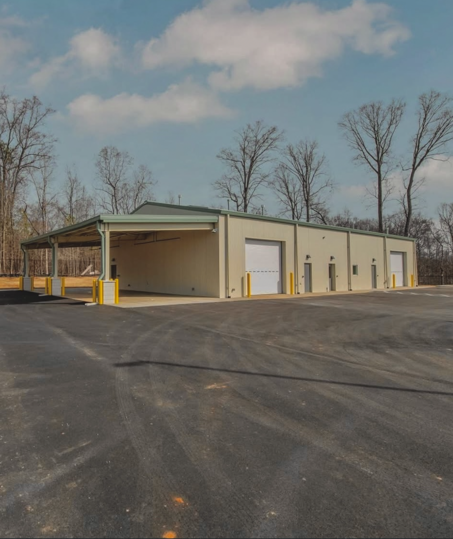 Exterior view of a beige metal storage or warehouse building with four garage doors, yellow and white traffic barriers, and a parking lot in the foreground. Bare trees and a partly cloudy sky are in the background.