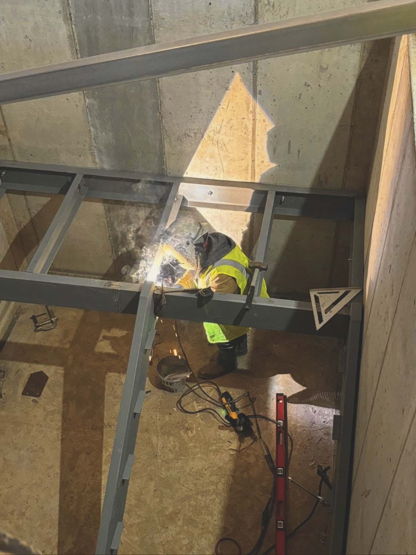 A construction worker in a yellow safety vest is welding inside a metal frame structure. Sparks and bright light are visible from the welding process, and various tools and equipment are scattered around on the floor.