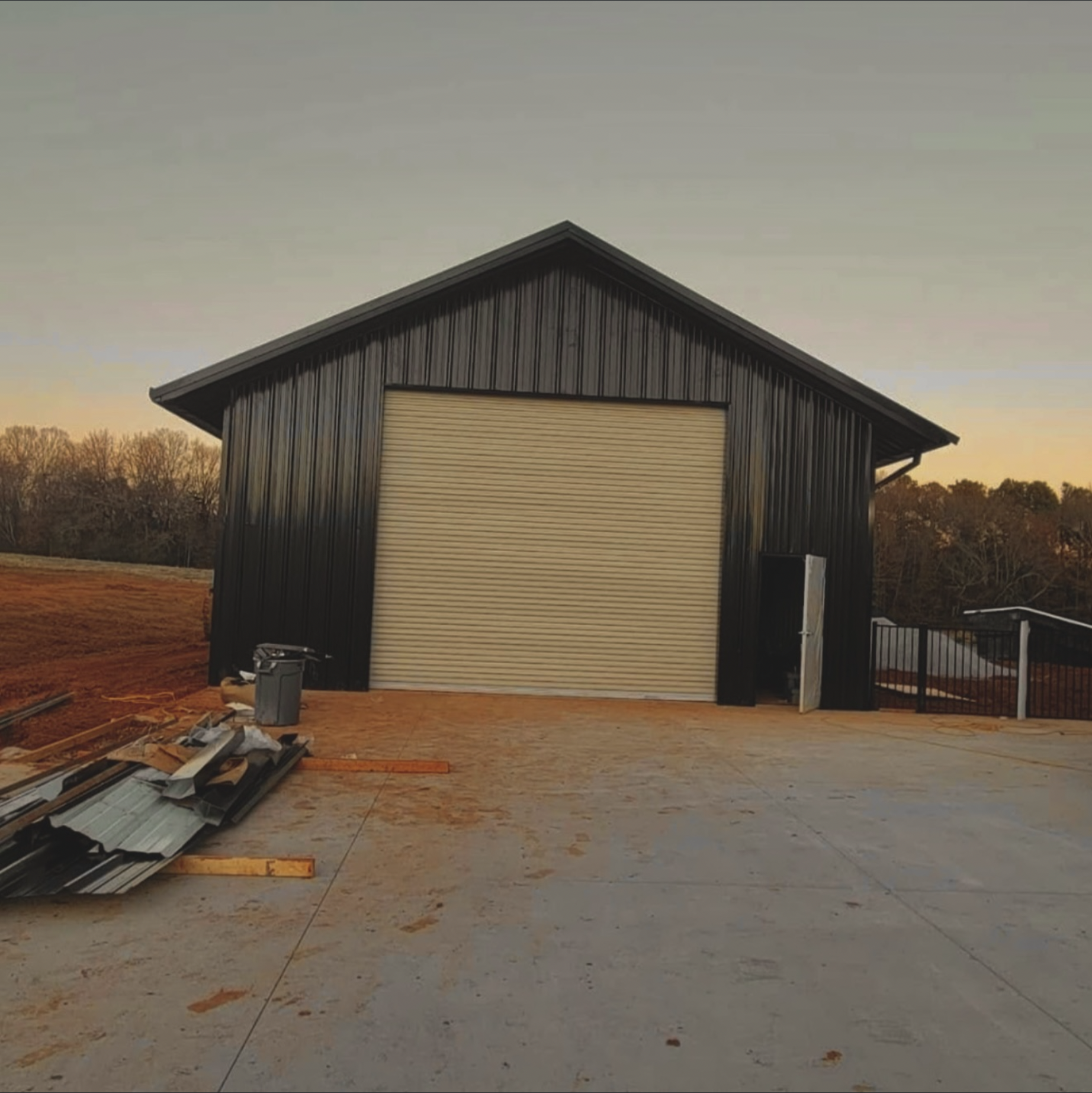 Construction site with partially built black metal building and a partially open garage door, scattered construction materials on the ground, and trees in the background during sunset.