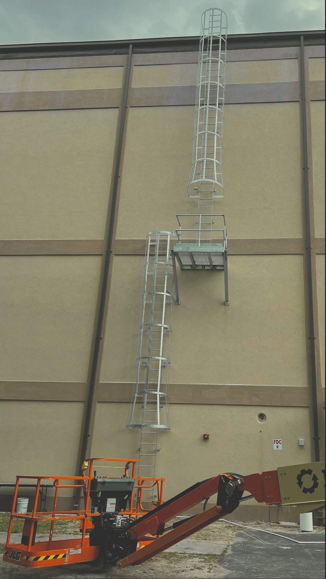 Construction workers are installing or repairing a tall outdoor fire escape ladder on a beige building with brown trim, using a cherry picker lift.