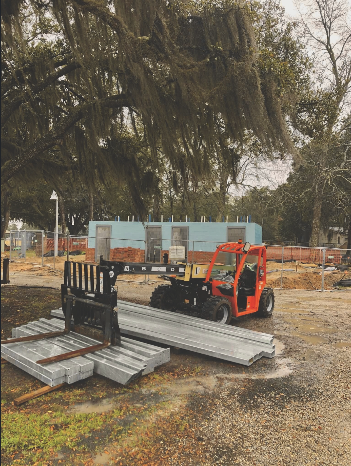 Construction site with metal beams, a small orange utility vehicle, a black bench, and a partially built blue structure behind a chain-link fence. Large trees with hanging moss are in the background.