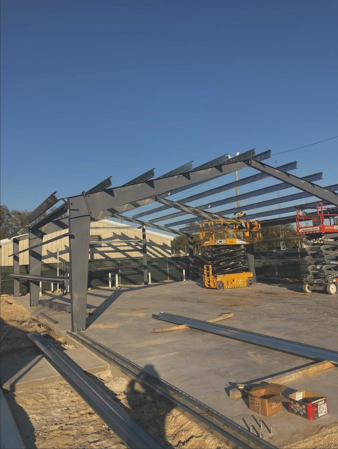 Construction site with metal framework for a building or structure, construction tools and equipment including a yellow lift, and a clear blue sky in background.