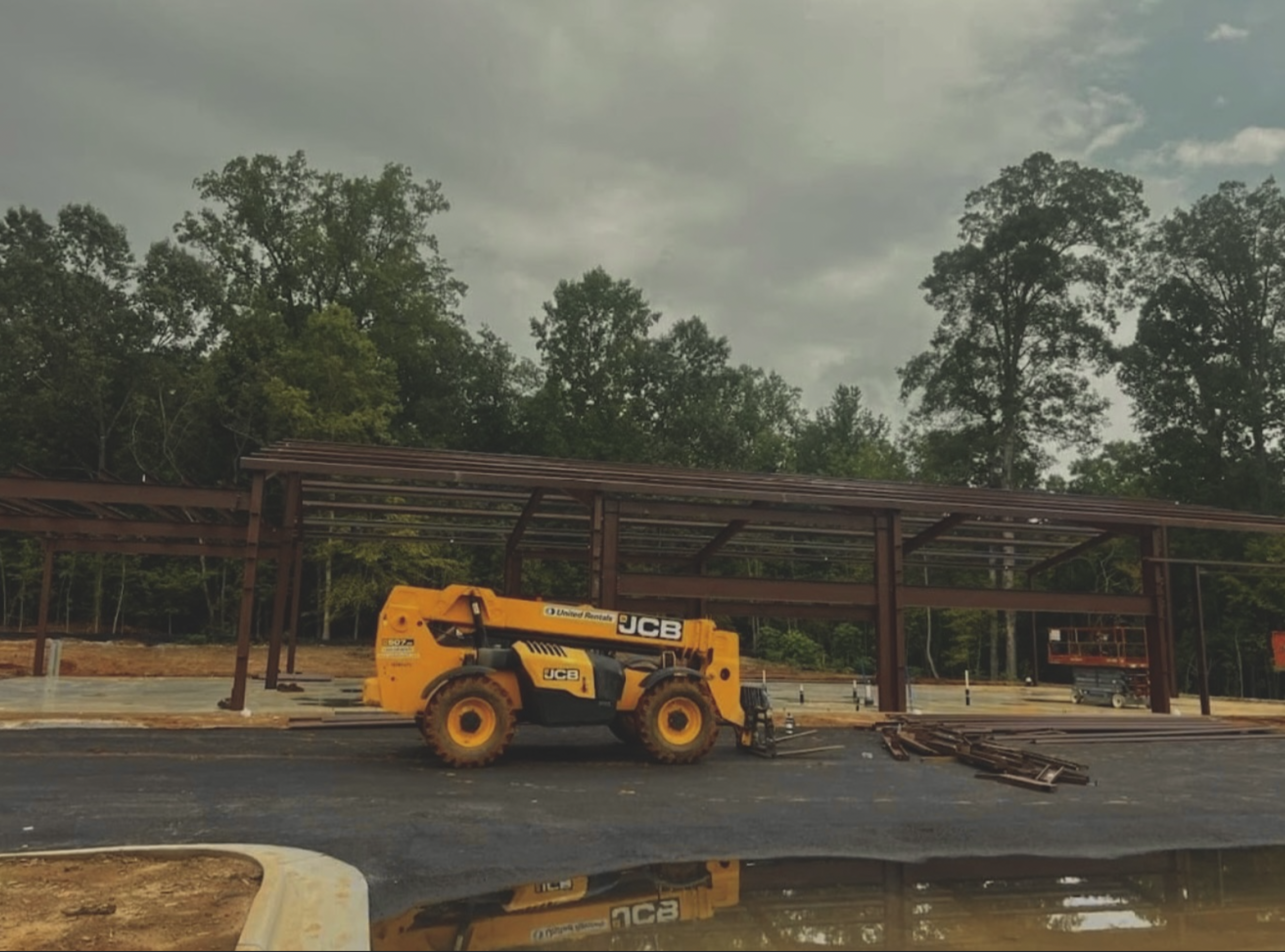 Construction site with steel framework for a building, a yellow JCB telescopic boom lift, and trees in the background under a cloudy sky.