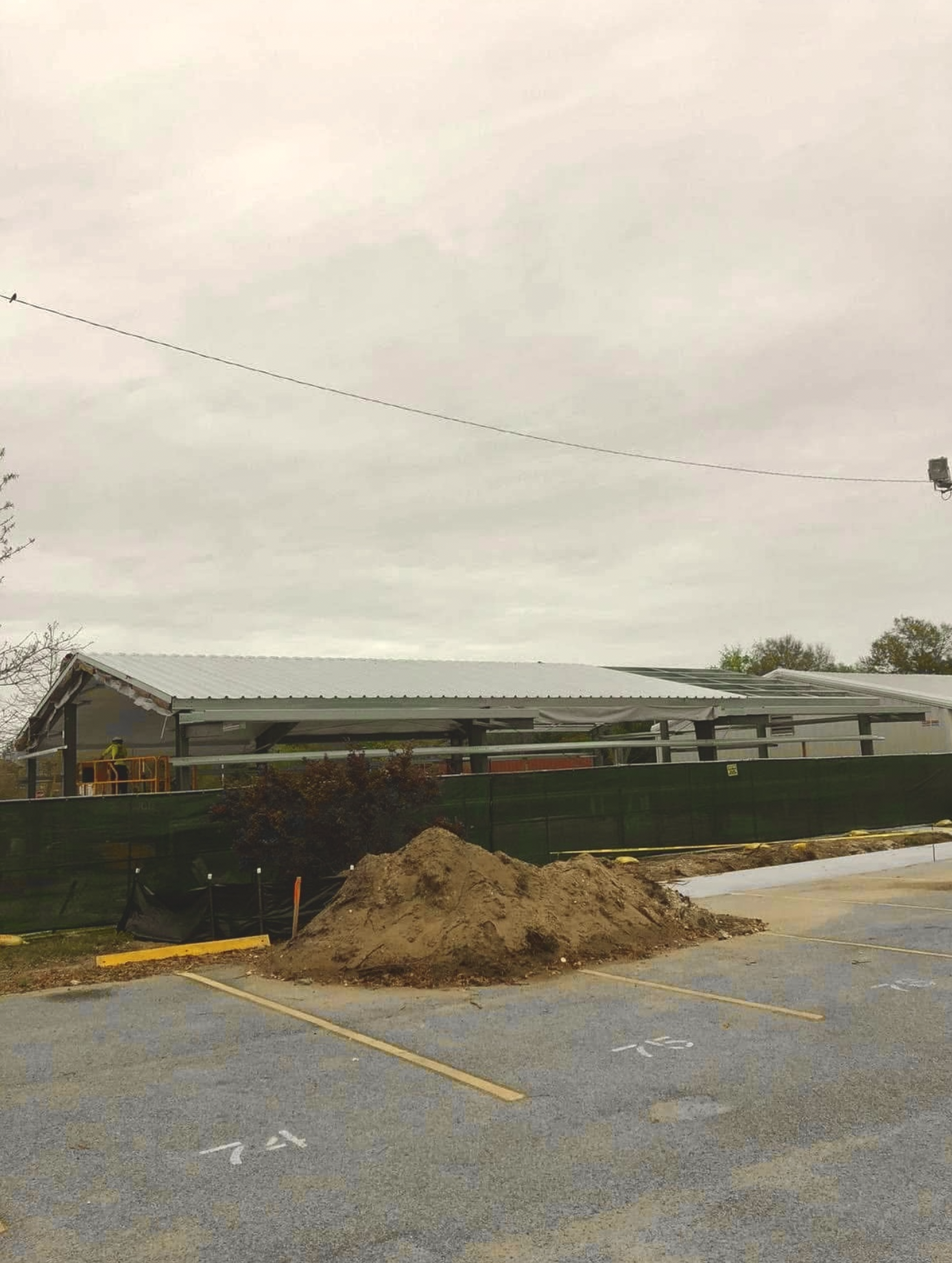 Construction site with a large mound of dirt, a green fence, and a building with a metal roof in the background. Overcast sky.