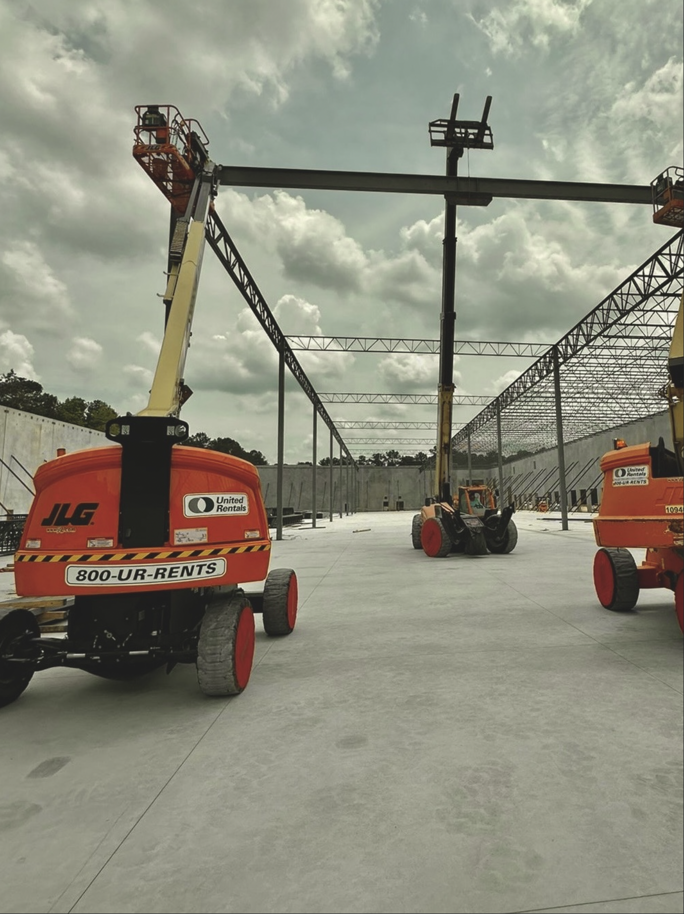 Construction site with large yellow scissor lift and a boom lift, metal framework being installed against a cloudy sky.