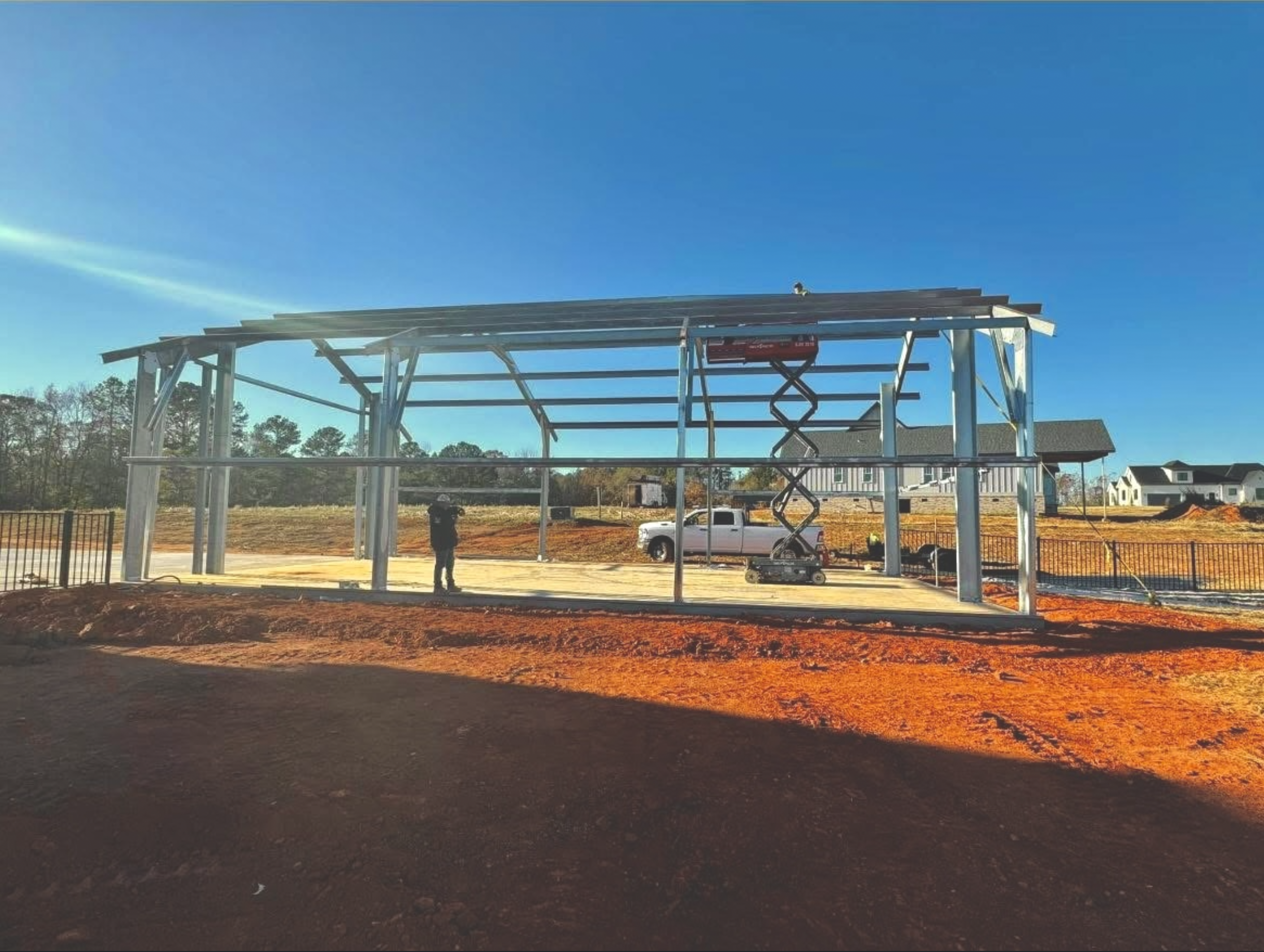 Construction site with a metal framework structure, construction worker, vehicle, wood pile and residential houses in the background under a clear blue sky.