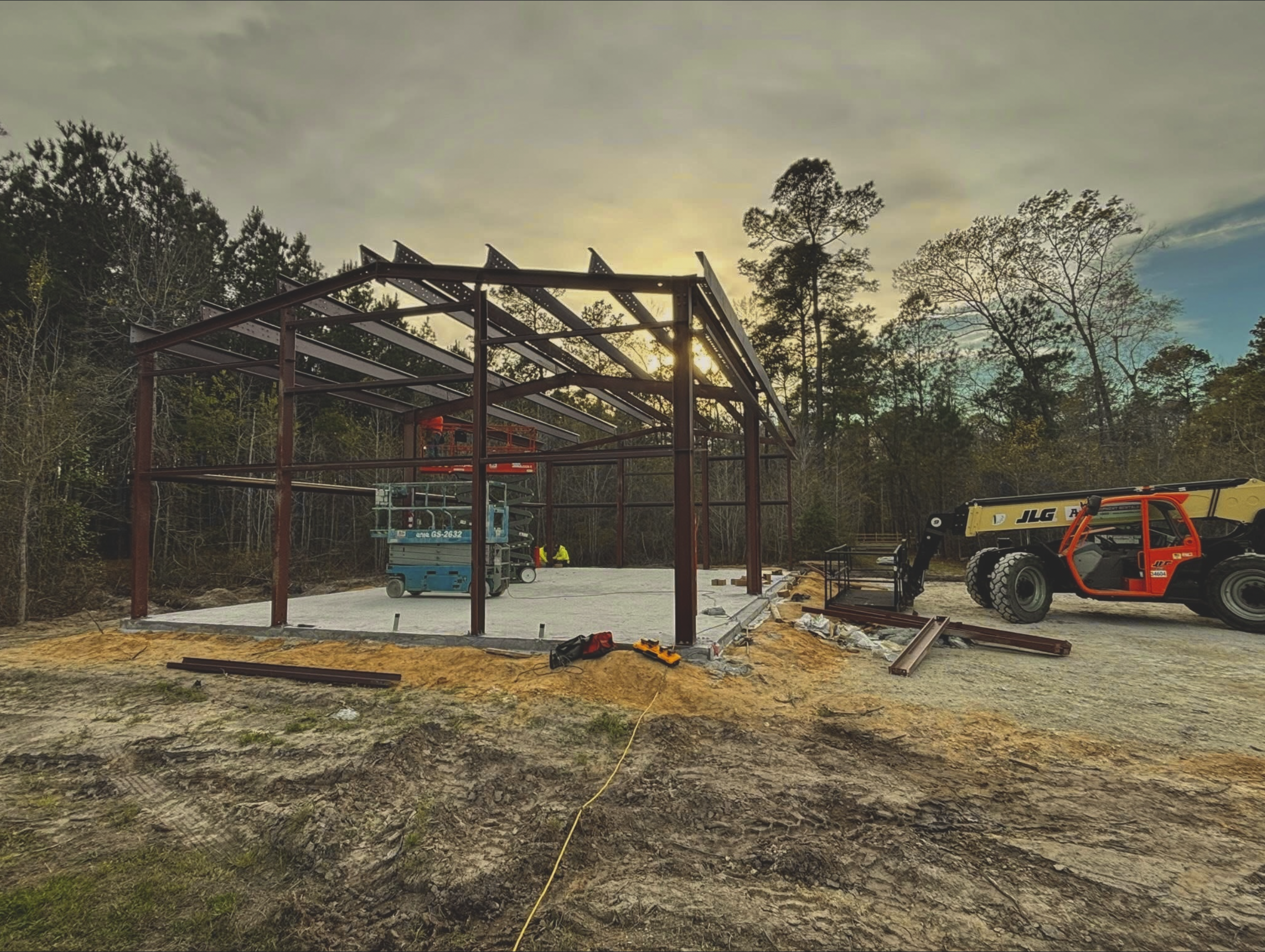 Construction site with steel framework for a building, a cherry picker lift, and construction equipment outdoors at sunset with trees in the background.