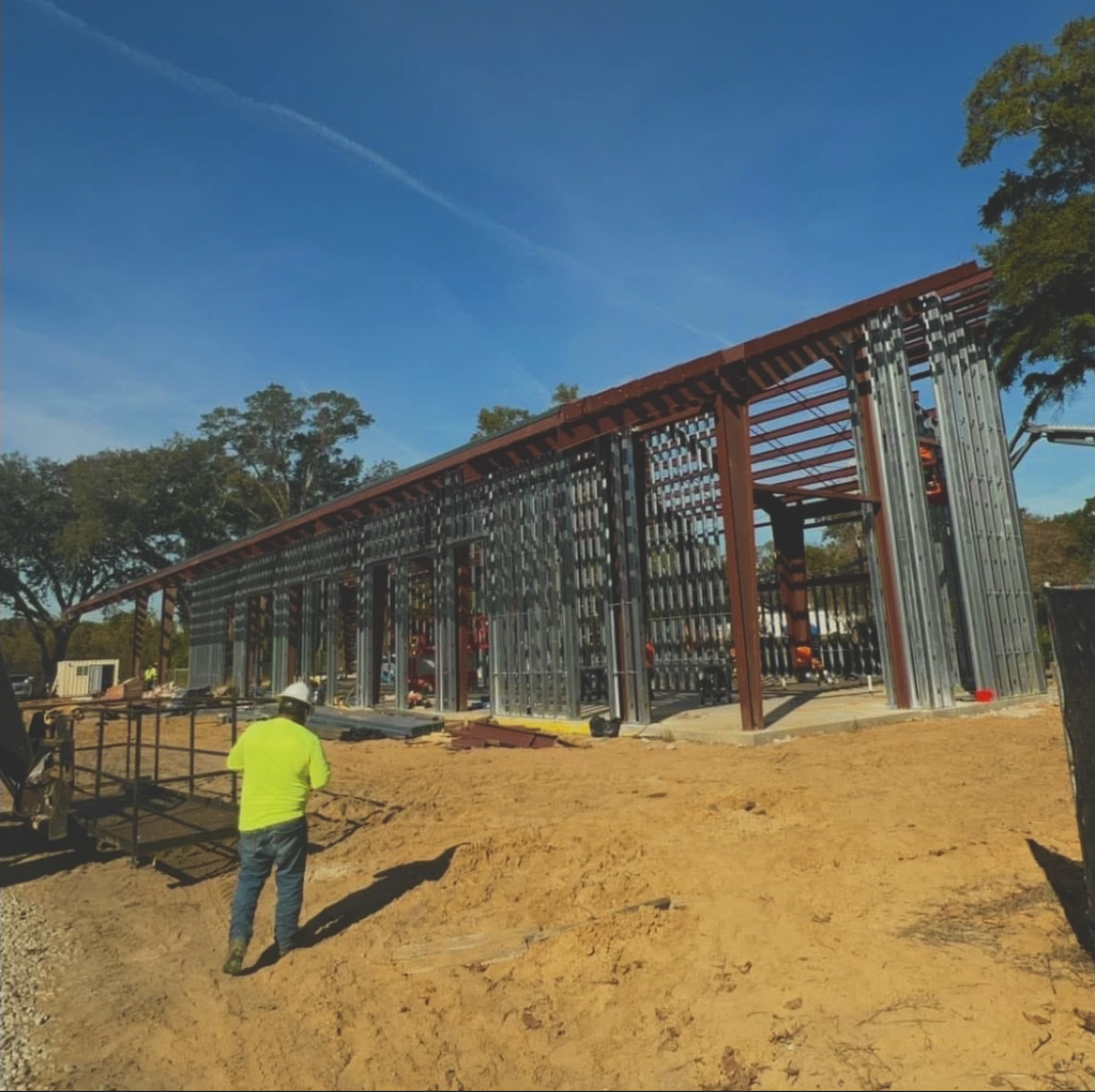 Construction site with a partially built steel structure, construction workers present, and a clear blue sky overhead.