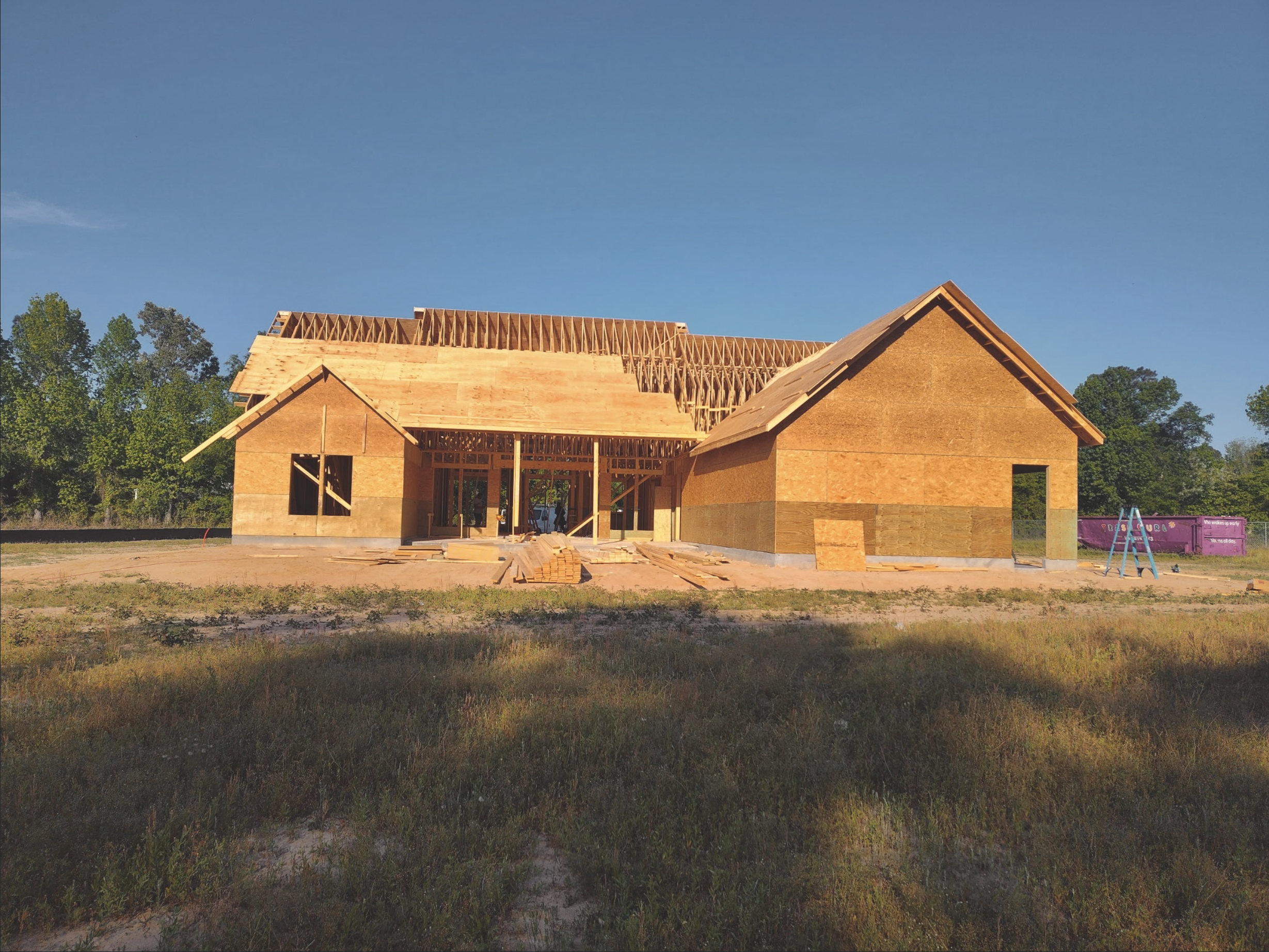 A house under construction with wooden framing and a roof, surrounded by trees and an open grassy area.