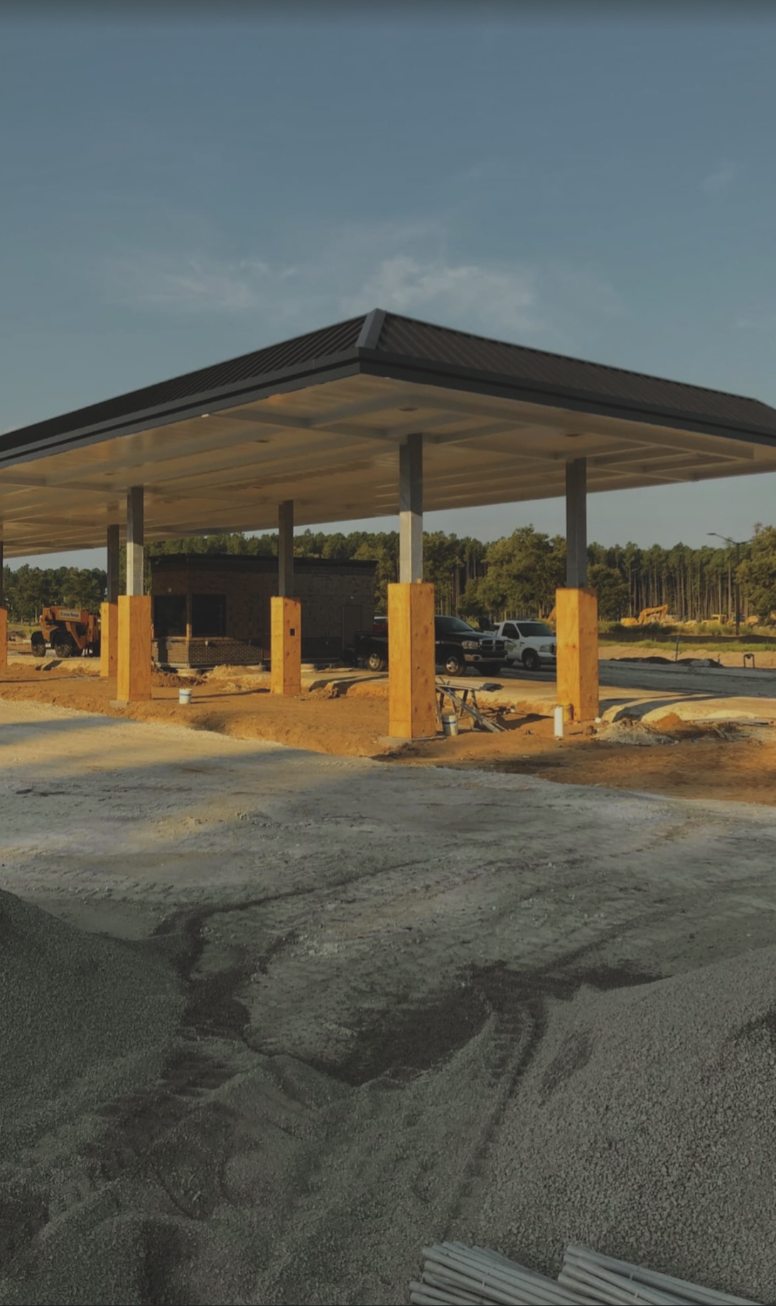 Construction site with a partially built canopy structure supported by wooden posts, with vehicles and construction equipment in the background, and a partly paved and dirt ground in the foreground.