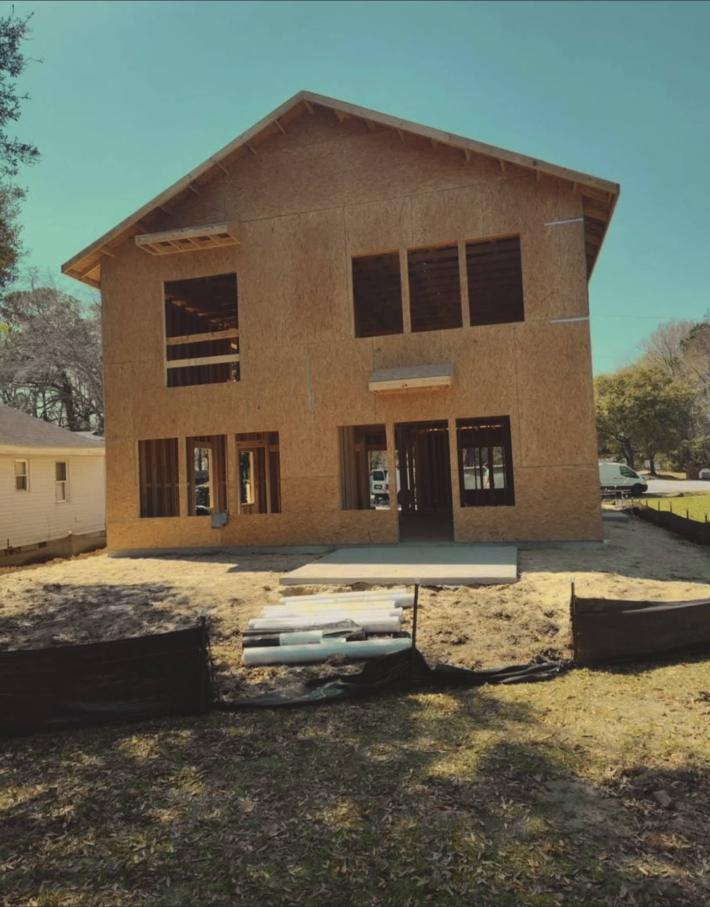 Unfinished two-story wooden house under construction with no exterior siding, windows, or doors installed, on a dirt yard with construction materials nearby, under a clear blue sky.