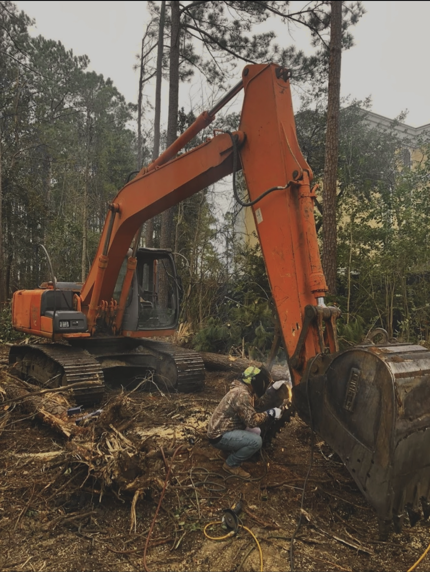 A person working outdoors in a forested area with a large orange excavator and fallen tree debris, wearing camouflage jacket, kneeling near the machinery.