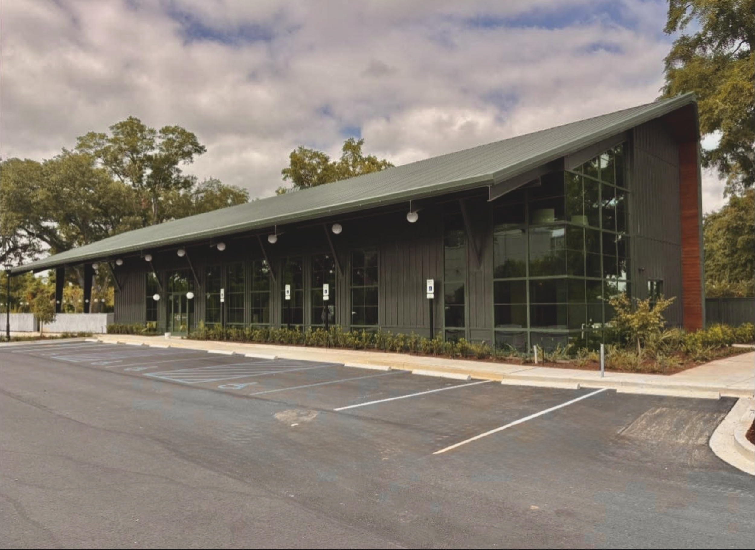 An empty parking lot in front of a modern building with large glass windows and a metal roof, surrounded by trees with autumn foliage and a partly cloudy sky.