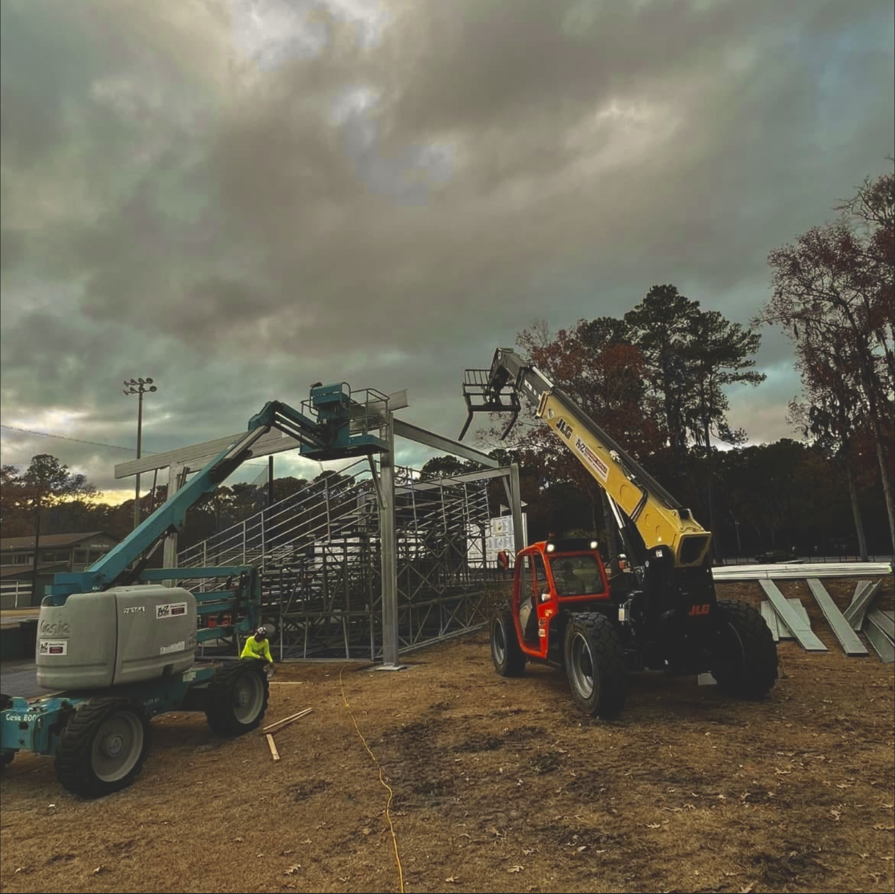 Construction site with lift equipment, a worker in a yellow safety vest, and a cloudy sky surrounded by trees.
