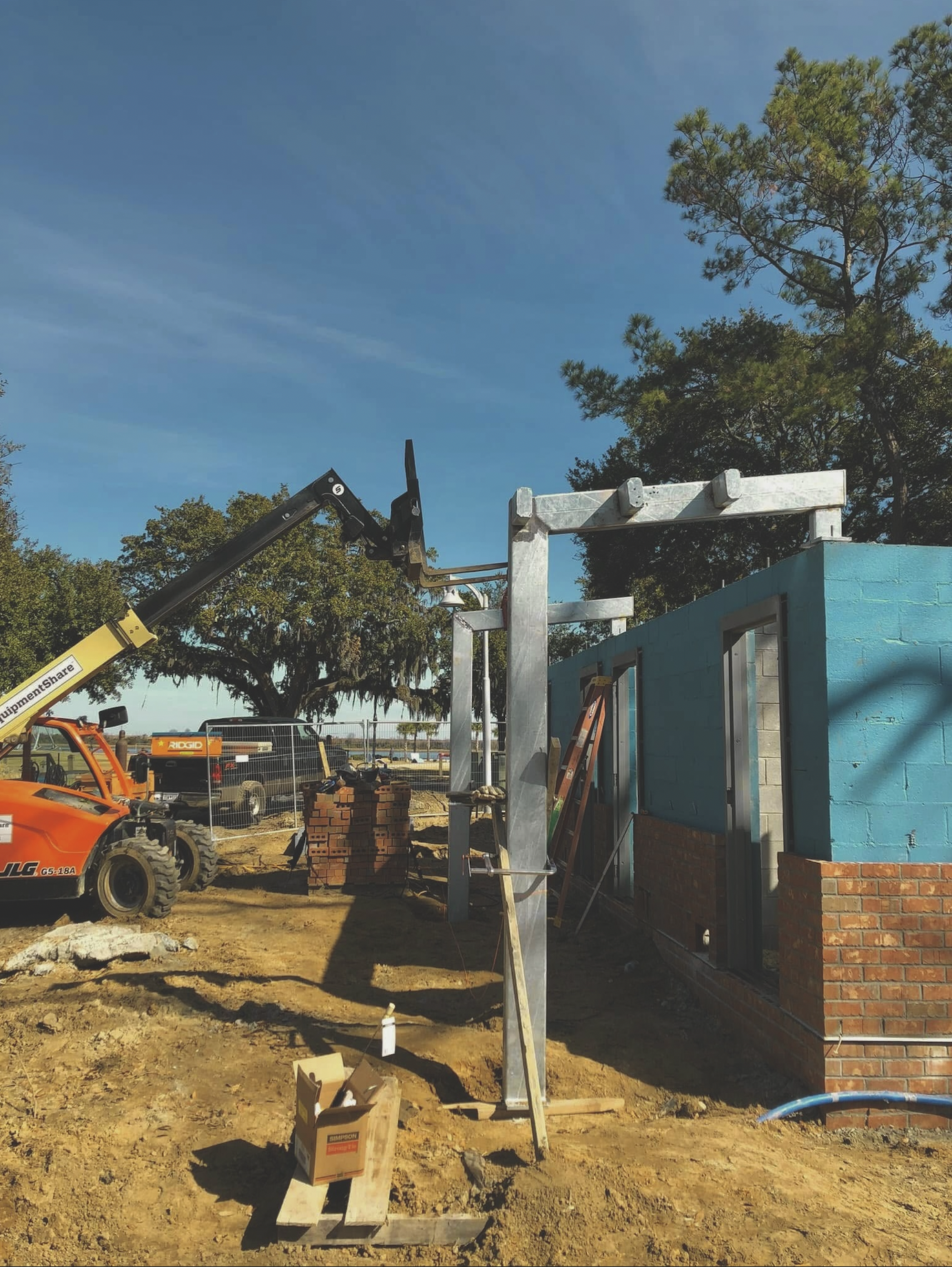 Building under construction with concrete block walls and steel framing. Construction equipment and materials on the site, with trees and a blue sky in the background.