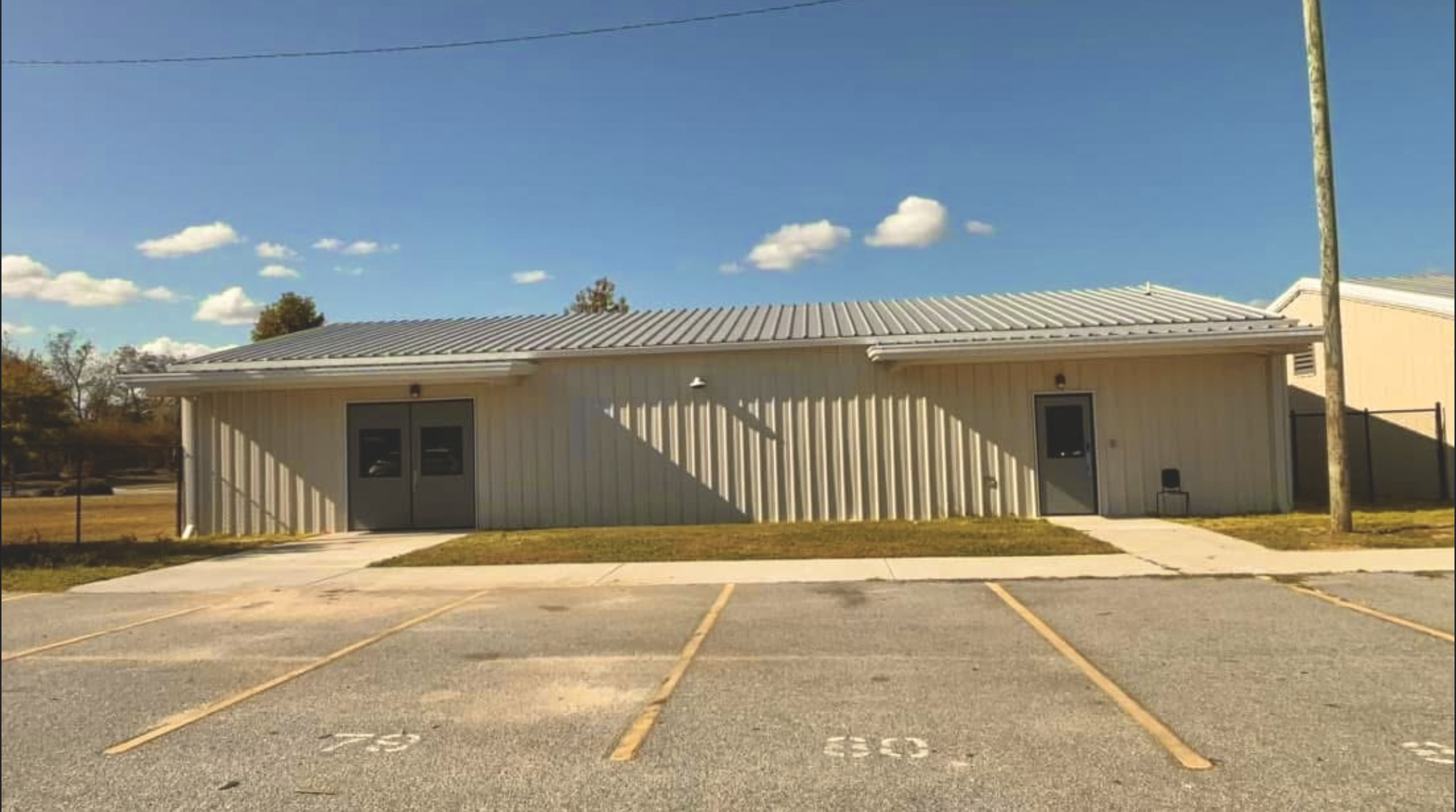 A single-story beige building with a metal roof and two doors, situated in a parking lot with some parking spaces marked. There is a sidewalk in front and a power pole to the right, with a background of trees and a partly cloudy sky.