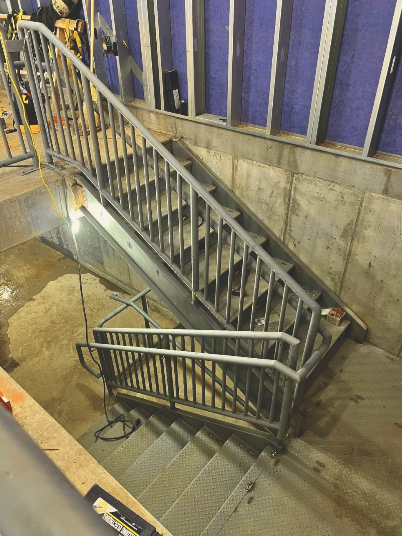 Construction area with a metal staircase and guardrails, blue wall panels, construction tools, and electrical cords.