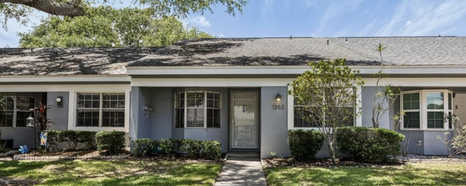Front view of a single-story house with a gray exterior, front door, and several windows, surrounded by a lawn and shrubs under a partly cloudy sky.