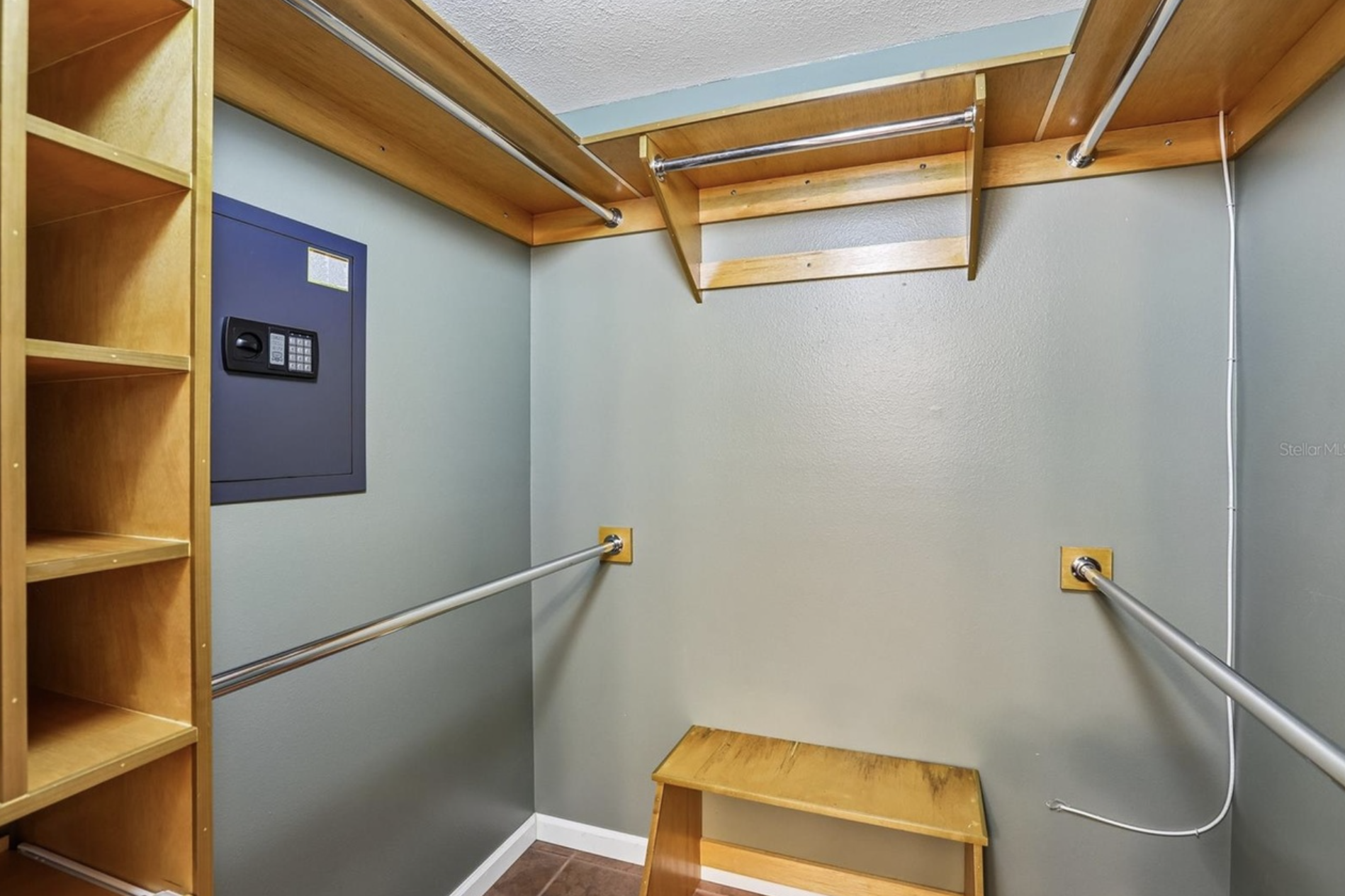 Walk-in closet with built-in wooden shelves, metal hanging rods, a safe embedded in the wall, and wooden bench.