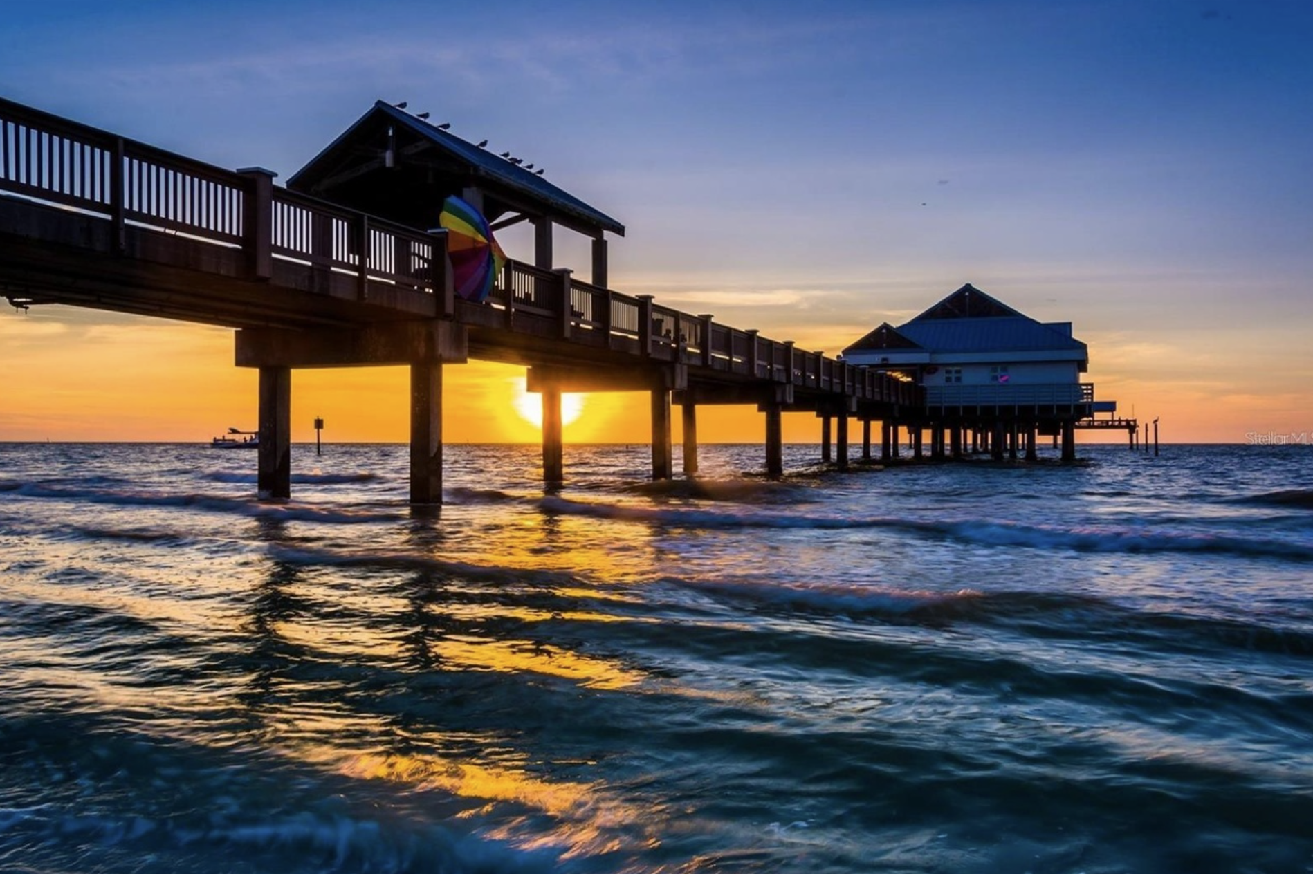 A pier extending over the ocean at sunset, with the sun partially visible behind the structure, a rainbow-colored umbrella hanging on the pier, and a boat in the distance.