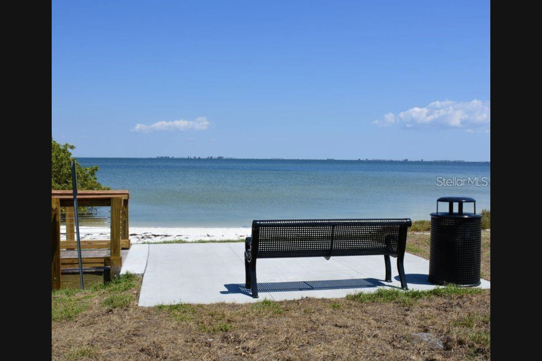 View of a beach with a clear blue sky, calm ocean, and a wooden deck with black chairs and a trash can in the foreground.