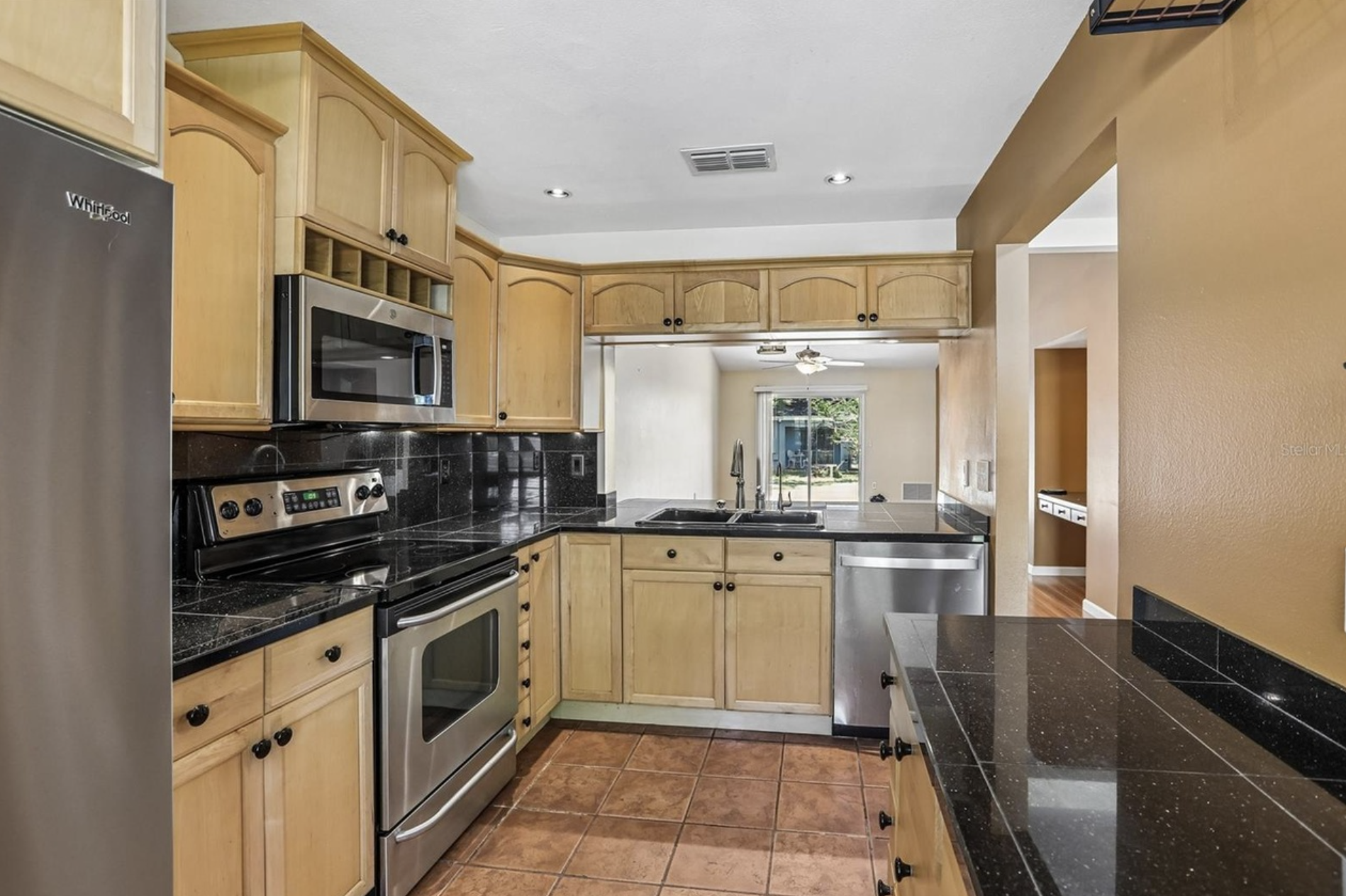 The kitchen has brown tiled floor, with daylight coming in through the front window and a view of a backyard through the open counter, with additional food prep area against the adjacent wall.