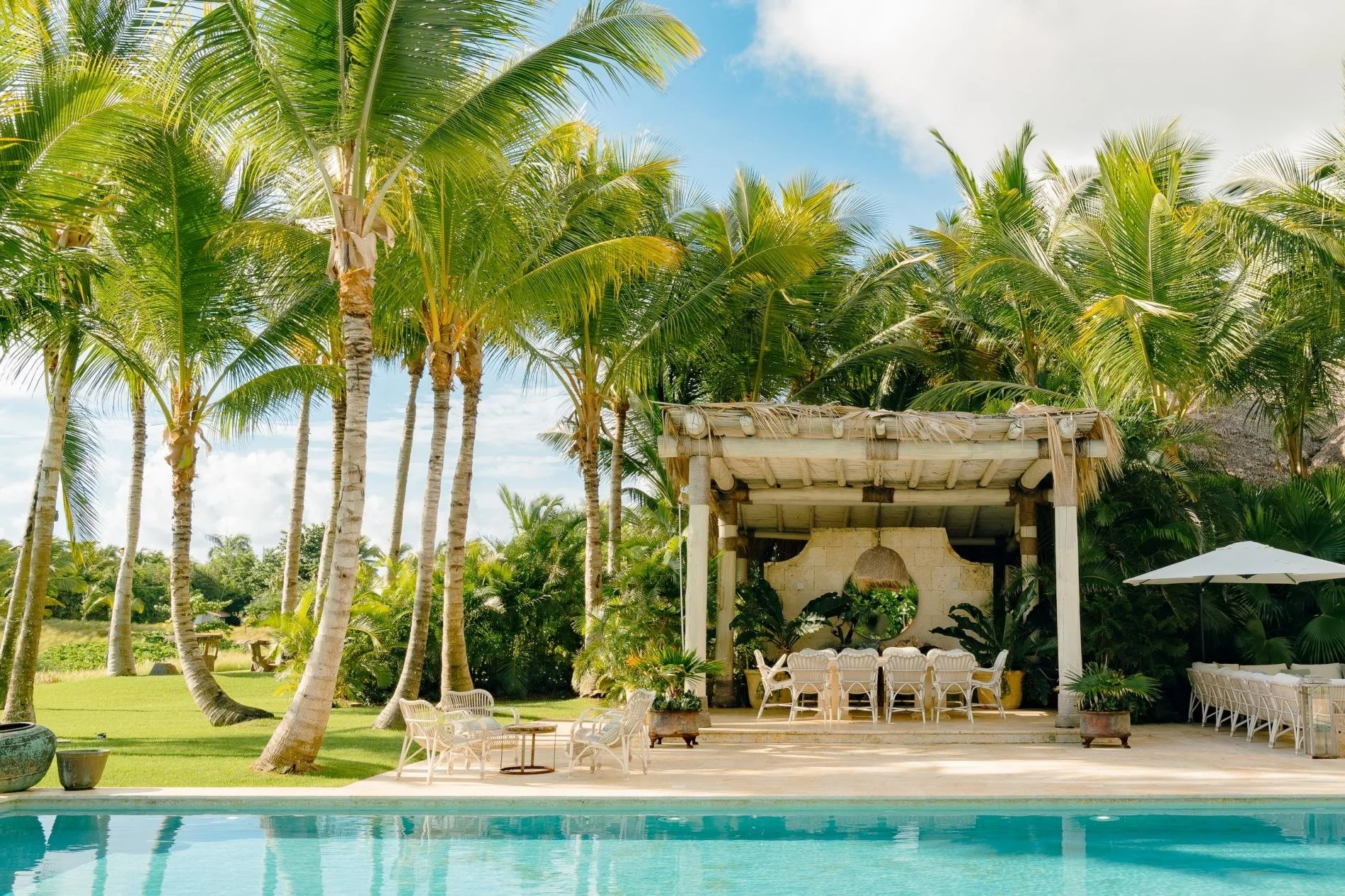 Swimming pool with clear blue water in a tropical setting surrounded by tall palm trees. There is a shaded outdoor seating area with white chairs and tables, and a large white umbrella on the right side. The sky is partly cloudy.
