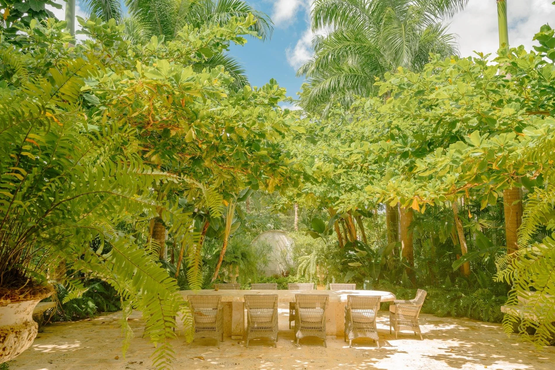 Outdoor seating area with six chairs around a stone table, surrounded by lush tropical greenery including palm trees and ferns under a partly cloudy sky.