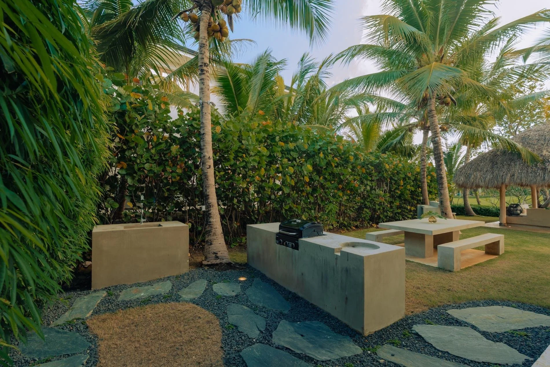 A tropical outdoor scene with palm trees, lush green bushes, a stone pathway, and a concrete seating area with a built-in barbecue and sink, under a thatched-roof umbrella.