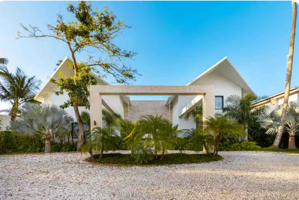 Modern beachfront house with white walls, palm trees, and a gravel driveway, under a blue sky.