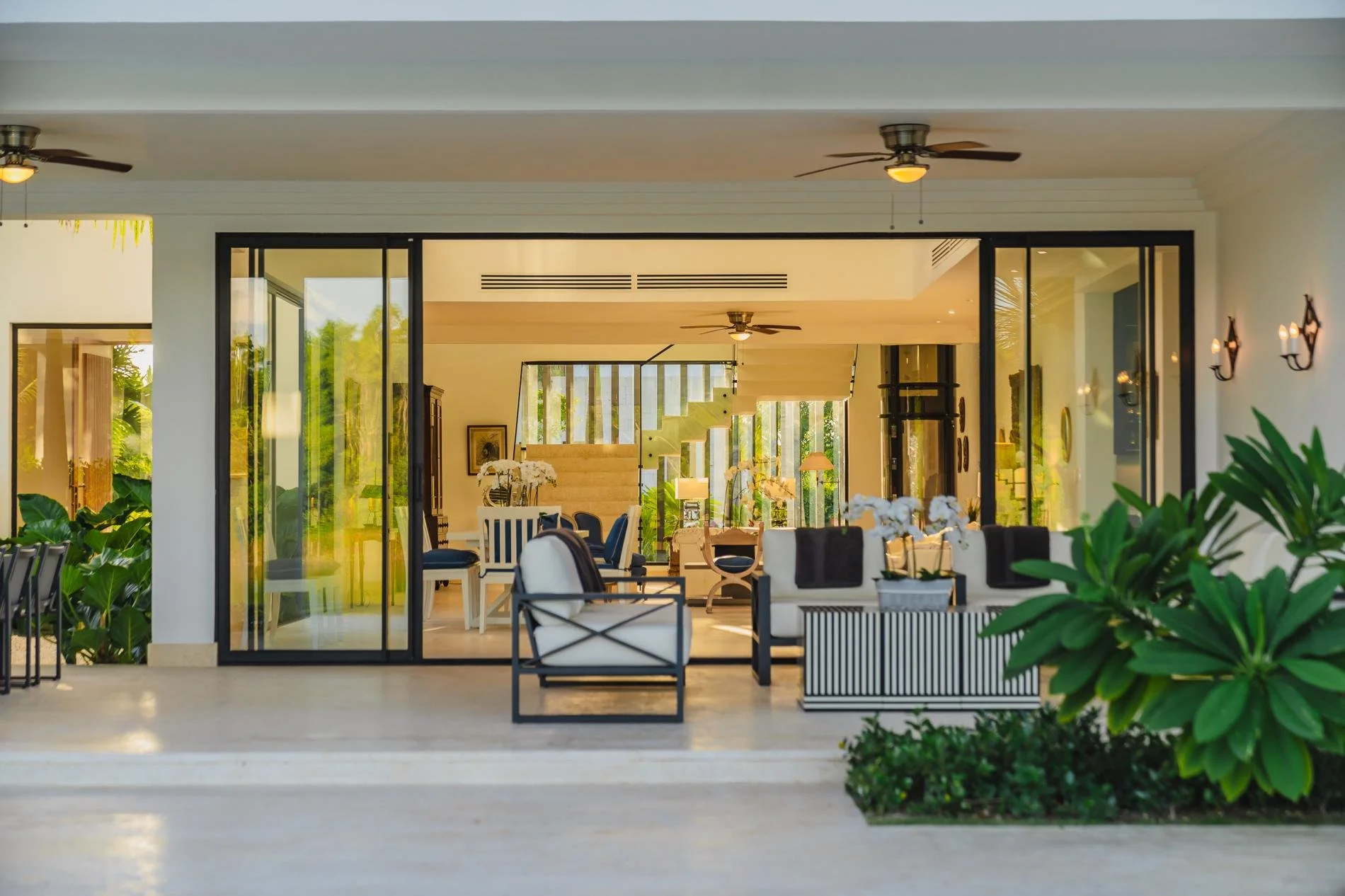 View of a modern indoor living space seen through a large sliding glass door, with white walls, ceiling fans, indoor plants, and furniture including chairs and a dining table.