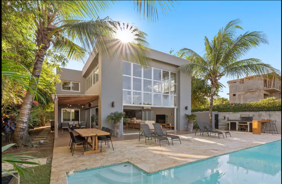 Modern two-story house with large glass windows, surrounded by palm trees, with a swimming pool and outdoor seating area on a sunny day