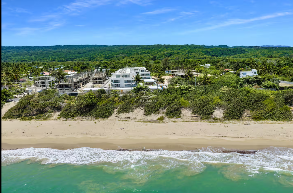 A beach with sand and waves in the foreground, vegetation and modern houses on a hillside in the middle ground, and a mountain or forested area in the background under a blue sky.