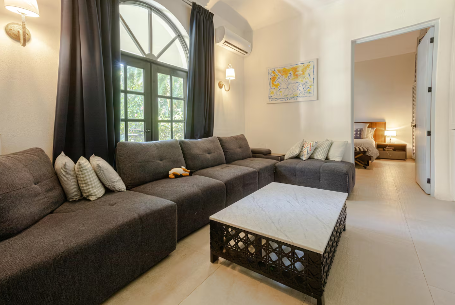 Living room with a dark gray sectional sofa, throw pillows, a marble-topped coffee table, large window with black curtains, and a doorway leading to a bedroom.