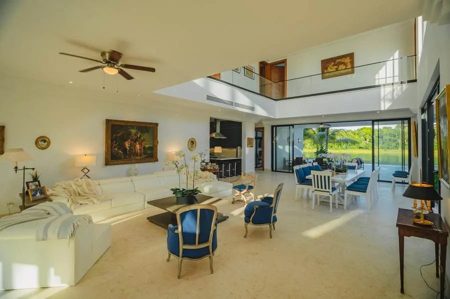 Spacious living room with white sofa, blue chairs, and a dining table, large glass sliding doors opening to greenery, and artwork on the walls.