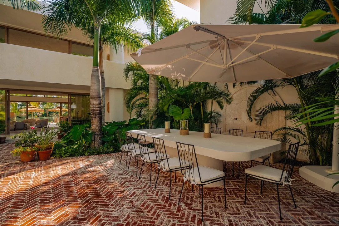 Outdoor dining area with a large white table, white cushioned chairs, a large white umbrella, tropical plants, and brick flooring.