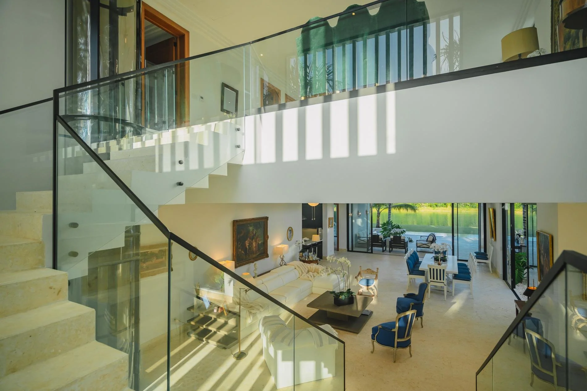 Interior view of a modern, spacious living room with large glass doors opening to an outdoor pool area. The room features a white sectional sofa, blue and gold armchairs, a fireplace, artwork on the walls, and a dining table set with chairs, with a view of green landscape outside.